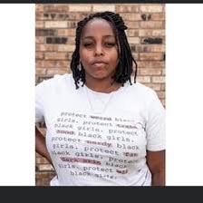 Image description: photo of Kyra Jay in front of a brick wall. Kyra wears a white t-shirt with text on it.